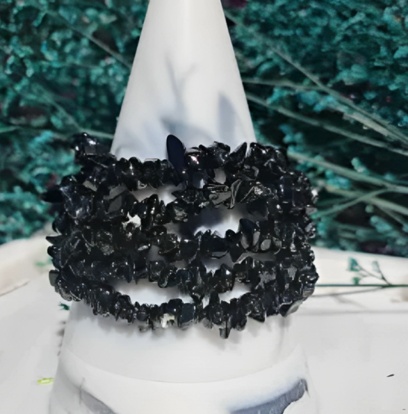 A black tourmaline chip bracelet displayed on a white stand against a backdrop of greenery, showcasing its protective and rugged design.