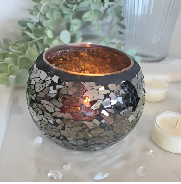 A black mirror crackle glass votive candle with a lit candle inside, placed on a white surface with a few scattered candle wicks and a plant in the background.